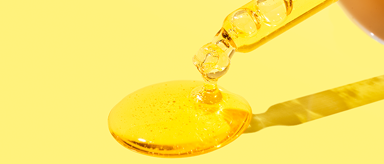 Closeup shot of serum dropper from Beekman 1802's Golden Booster Vitamin c & amla berry Brightening Serum, dropping orange serum onto the yellow ground. 