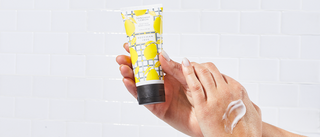 Up close shot of hand holding a tube of Beekman 1802's Sunshine Lemon Hand Cream, with other hand propped up showing the cream applied on the back of the hand, on a white tile background.