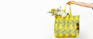 Extended hand holding out Beekman 1802's Limited Edition Baby Goat Tote, which has cartoon goaties face printed in a pattern on the tote, and the bag is filled with flowers and sunglasses hanging off the side on a white background.  