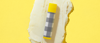 Small Tube of Beekman 1802's Pure Goat Milk Lip Balm in gingham packaging with a yellow cap, on top of lip balm ingredient smear on a yellow background. 