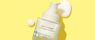 Bottle of Beekman 1802's Milk Primer SPF Sunscreen Primer on a yellow background with Milk Primer drops surrounding the bottle. 