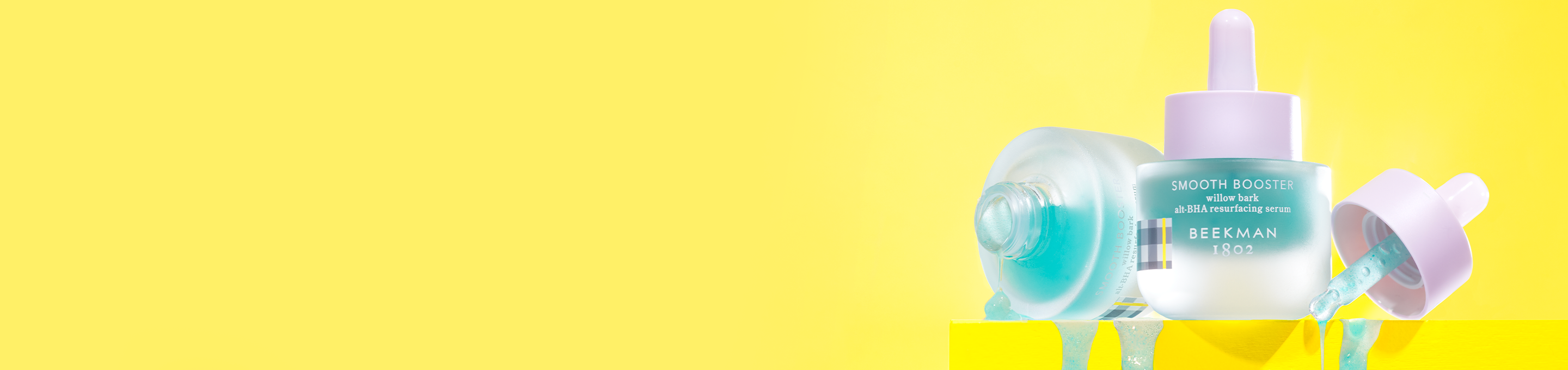 2 bottles of Beekman 1802's Smooth Booster Willow Bar Alt-BHA Resurfacing Serum on the far right of the screen, with one bottle standing up right in between the other bottle, which is unscrewed on the left, and the dropper on the right, with blue serum dripping over yellow ledge, on a yellow background.