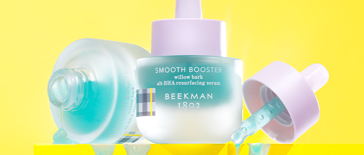 2 bottles of Beekman 1802's Smooth Booster Willow Bar Alt-BHA Resurfacing Serum, with one bottle standing up right in between the other bottle, which is unscrewed on the left, and the dropper on the right, with blue serum dripping over yellow ledge, on a yellow background.