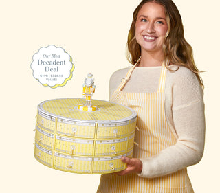 Image of model holding Beekman 1802's 2023 Slice of Kindness Advent Calendar and looking at the camera smiling while wearing a yellow and white striped apron on a cream colored background, with the words "Our Most Decadent Deal, $175 and a $320.50 value!"