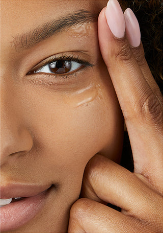 Up close shot of half of models face while she is looking into the camera and smiling, while using her fingers to apply the Beekman 1802 Dewy Eyed Illuminating Serum under her eyes. 
