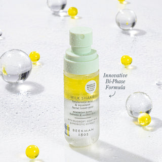 Bottle of Beekman 1802's Mini Milk Shake Toner Mist surrounded by clear and yellow spheres with the words "innovative Bi-Phase Formula" next to the bottle.