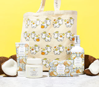 Image of Beekman 1802's Fall Seasonal Bounty Subscription Tote which shows the whipped body cream, hand cream, bar soap, and hand & body wash in front of a tote bag that is covered with a pattern of a cartoon goatie next to a pumpkin, surrounded by broken coconut pieces on a yellow background.