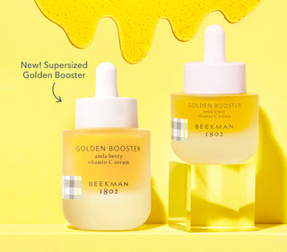 Bottle of Beekman 1802's Supersized Golden Booster Serum positioned right next to a bottle of regular sized golden booster serum thats on an acrylic riser right, both on a yellow background with serum drips on the background and with the products next to the words "New! Supersized Golden Booster" and an arrow pointing to serums.