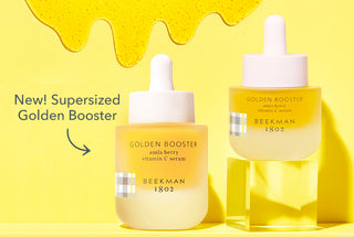 Bottle of Beekman 1802's Supersized Golden Booster Serum positioned right next to a bottle of regular sized golden booster serum thats on an acrylic riser right, both on a yellow background with serum drips on the background and with the products next to the words "New! Supersized Golden Booster" and an arrow pointing to serums.