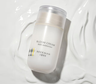 Bottle of Beekman 1802's Bloom Cream Daily Moisturizer laying flat on the moisturizer cream itself thats colored white