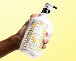 Hand model holding Lavender Hand & Body wash with foamy soap around it on a yellow background