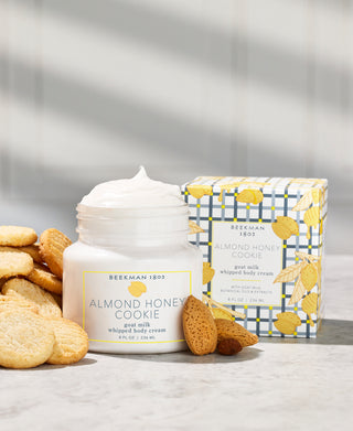 Jar of Beekman 1802's Almond Honey Cookie Whipped Body Cream opened and surrounded by cookies, with the packaging box in the background.