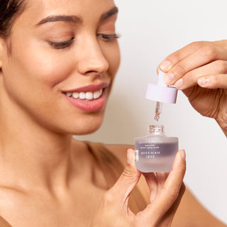 Up close shot of model holding a bottle of Beekman 1802's Dream Booster Bakuchiol better aging serum with one hand, and holding the dropper up in the other hand, while looking down and smiling, on a white background.
