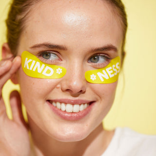 Headshot of model wearing Beekman 1802's yellow Kindness Undereye Mask under her eyes while looking away from the camera and smiling and her hand touching the side of her face, all on a yellow background.
