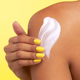 Close up image of models shoulder while they are using their hand with yellow nail polish to rub Beekman 1802's Ceramide Goat Milk Whipped Body Cream onto their skin on a yellow background.