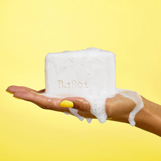 Up close shot of hand holding a wet, sudsy bar of Beekman 1802's Jasmine Bloom Goat Milk Soap on a yellow background.