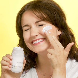 An image of a model holding the Beekman 1802 Bloom Cream Daily Moisturizer with one hand and applying the cream with the other hand on the side of her face while smiling.