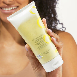 An image of a model holding the beekman 1802 sunshine exfoliating scrub up close while smiling in the background.