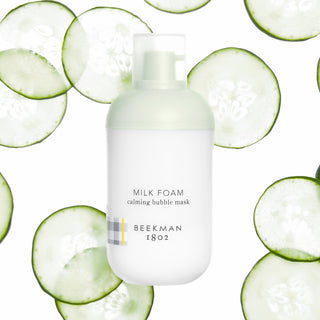 Bottle of Beekman 1802's Milk Foam Calming Bubble Mask on a white background, surrounded by thinly sliced cucumbers. 
