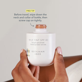 Up close shot of hand holding uncapped bottle Beekman 1802 Milk Tint SPF 43 Tinted Primer Serum, with an arrow pointing to the top saying "pro tip: Before travel, wipe fown the neck and collar of bottle, then screw cap on tightly."