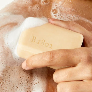 Up close shot of hand holding a wet bar of Beekman 1802's Vanilla Woods Palm-Sized Goat Milk Soap that's covered in suds while rubbing bar onto their shoulder.