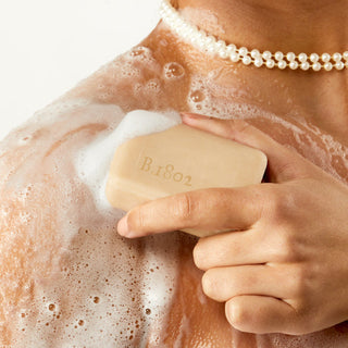 Up close shot of models hand holding a wet bar of Beekman 1802's Ylang Ylang & Tuberose Palm-Sized Goat Milk Soap that's covered in suds while rubbing bar onto their shoulder and wearing a pearl necklace.