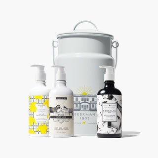 Bottles of Beekman 1802's Pure, sunshine Lemon and vanilla absolute goat milk lotions all lined up next to each other in that order, standing in front of the grey milk pail tin that says "beekman 1802" on the front, all on a light gray background. 