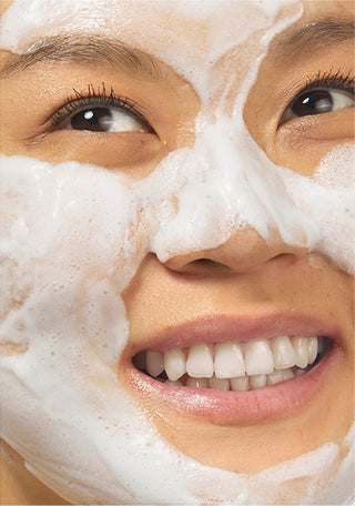 Up close shot of model smiling with her teeth and looking off to the right, while wearing the Beekman 1802 Milk Foam calming Bubble mask on her face, showing all the bubbles that have formed on her skin. 