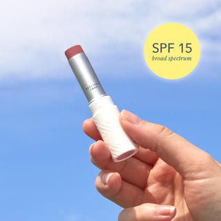 Upclose shot of hand holding Beekman 1802's Rosy Posy SPF 15 Goat Milk Tinted Lip Cream with sky in the background and a yellow bubble to the right of the product that says "SPF 15 Broad Spectrum"