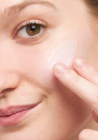 Up close shot of side of model's face while they are smiling and looking at the camera and applying the Beekman 1802 Milk Primer SPF serum onto the side of their face with their fingers. 