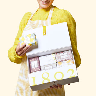 Image of model wearing a yellow sweater and apron while holding Beekman 1802's The G.O.A.T. Soap Set of 8 holiday gift box with one hand, which is opened revealing eight bar soaps inside, and holding one the bar soaps in the other hand, on a cream colored background.