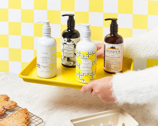 Two hands holding a yellow tray that has Bottles of Beekman 1802's Hand & Body washes, which include pure, vanilla absolute, sunshine lemon, and honey & orange blossom in that order, with baked cookies on the bottom left of the image, and on a yellow and white checkered background.