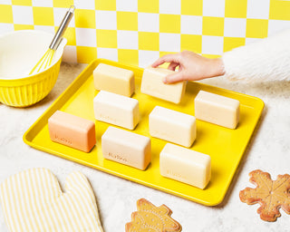 8 unwrapped Beekman 1802 goat milk bar soaps all lined up on a yellow baking tray, with a hand coming from the right side of the image picking up a bar soap, with the tray surrounded by cookies, oven mitts, a mixing bowl and whisk, on a yellow and white checkered background.