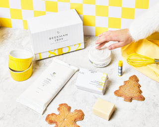Products from The Original Recipe Pure Goat Milk Gift Set next to gift box on a counter top, with a hand coming from the right side dipping into the whipped body cream, surrounded by cookies and whisks, and on a yellow and white checkered background.