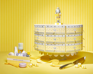 Full shot of Beekman 1802's 2023 Slice of Kindness Advent Calendar on a yellow striped background, with one of the drawers pulled completely out of the calendar filled with a bunch of Beekman 1802 skin and body products and placed to the right of the calendar, surrounded by candy and a yellow ribbon, and a cake slicer to the left of the calendar.