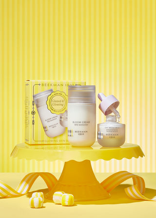 Bottle of Beekman 1802's Bloom Cream Daily Moisturizer and bottle of the oh mega milk serum standing next to each other on a yellow cake stand, with the dropper coming out of the serum bottle, and in front of the yellow gift box the Glazed & Glowing Advanced Hydration Gift set comes in, surrounded by yellow candy and on a white and yellow striped background.