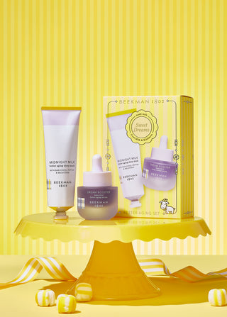 Tube of Beekman 1802's Midnight Milk Sleep Mask, and bottle of the Dream Booster better aging serum, standing next to each other on a yellow cake stand, in front of the gift box it comes in thats also on the cake stand, with pieces of yellow and white candy on the table underneath the stand, and on a yellow and white striped background.