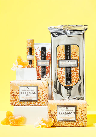 Products from Beekman 1802's Honey & Orange Blossom line all displayed next to each other one white acrylic risers, surrounded by sliced oranges and on a yellow background.