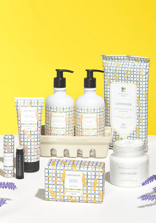 Products from Beekman 1802's Lavender Collection which includes the caddy set, lip balm, bar soap, body cream, face wipes, and hand cream all displayed next to each other, surrounded by purple lavender flowers on a yellow background. 