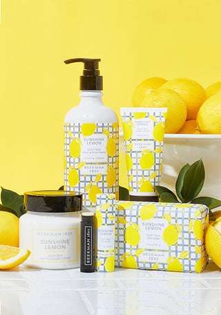 Products from Beekman 1802's Sunshine Lemon Line in front of a bowl of lemons and surrounded by sliced lemons, standing on white tile and on a yellow background. 