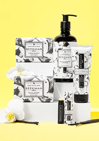 Products from Beekman 1802's Vanilla Absolute line all displayed next to each other on a white acrylic display risers, surrounded by vanilla ingredient and flowers on a white background.