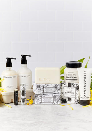 Products from Beekman 1802's Ylang Ylang & Tuberose collection which includes the caddy set, lip balm, bar soaps, body cream, and hand cream all displayed next to each other, surrounded by Ylang Ylang flowers and on a light grey tile background.  
