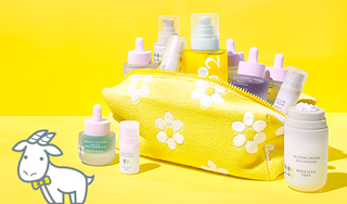 Image of a Yellow Dopp Bag covered in white flowers, filled with Beekman 1802's Clinically Kind Skincare and the Bloom Cream Daily Moisturizer with some serums standing around the bag, next to a white cartoon goatie near the serums on the left on a yellow background. 