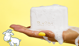 Closeup shot of hand with yellow nail polish holding a Beekman 1802 Sudsy Bar Soap, with the words "B.1802" engraved in the soap, and next to an image of a white cartoon goatie on a yellow background.