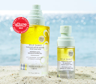 Bottle of Beekman 1802's Milk Shake Toner Mist next to a mini bottle of the Milk Shake toner mist on the sand at the beach, with an Allure "Best of Beauty Award Winner" Badge on the top left. 
