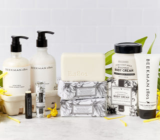 Collection shot of the Beekman 1802's full-line of Ylang Ylang & Tuberose Bodycare Products which includes the bar soap, body cream, hand cream, lip balm, hand and body wash, lotion, and caddy, all stacked up next to each other surrounded by yellow flowers and on a grey tile background.  
