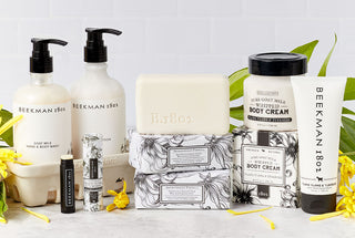 Collection shot of the Beekman 1802's full-line of Ylang Ylang & Tuberose Bodycare Products which includes the bar soap, body cream, hand cream, lip balm, hand and body wash, lotion, and caddy, all stacked up next to each other surrounded by yellow flowers and on a grey tile background.  