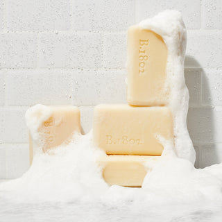 An image of 4 sudsy unwrapped Beekman 1802 9oz Coconut Cream Bar Soaps stacked next to each other surrounded b suds on a white tile background.