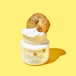 Opened jar of Beekman 1802's Fresh Baked Cookies & Milk Whipped Body Cream, with a chocolate chip cookie inserted on top of the whipped body cream product, on a yellow background.