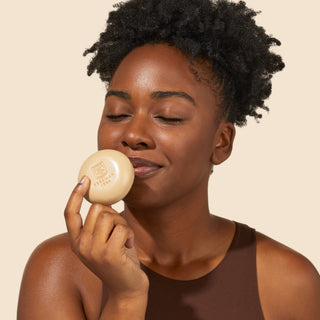 Shot of model holding one of the goat milk soaps from Beekman 1802's Fresh Baked Cookies & Milk Round Bar Soap Trio up to her nose, while her eyes are closed and smelling the bar soap, on a cream colored background.